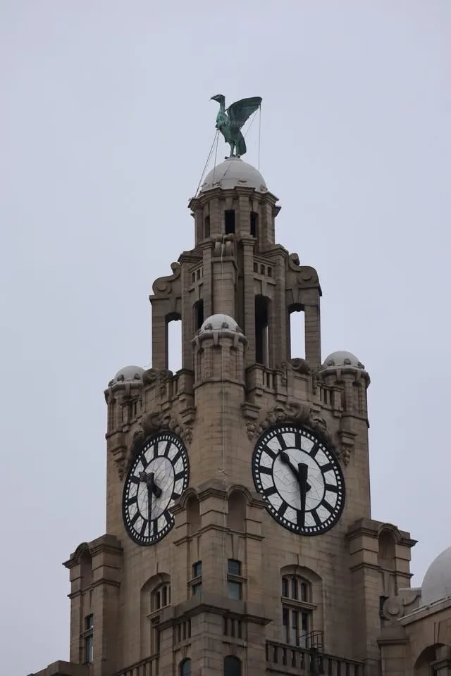 A tall stone clock tower with two large clock faces, topped by a green statue of a bird with outstretched wings. The…