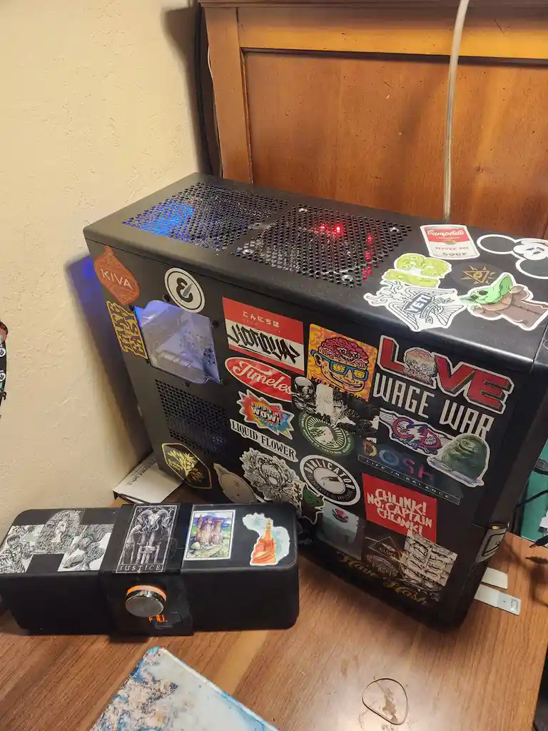 A black computer case covered in numerous colorful stickers, including 'Love Wage War', 'Campbell's Pepper Pot Soup'…