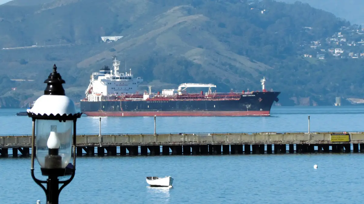 A large black and red cargo ship named 'WEST VIRGINIA' sails on blue water with green hills in the background. In the…