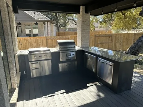 An outdoor kitchen setup under a covered patio features a stainless steel grill, sink, cabinets, and dishwasher, all set on…
