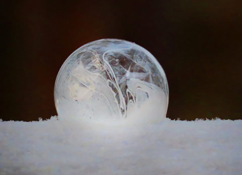A clear, spherical ice globe with internal swirling patterns rests on a bed of white snow against a dark brown background…