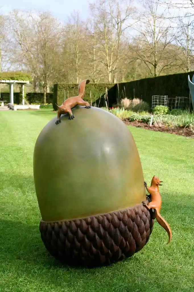 A large bronze sculpture of an acorn sits on a lawn, with two small bronze squirrel figures—one perched on top and one…