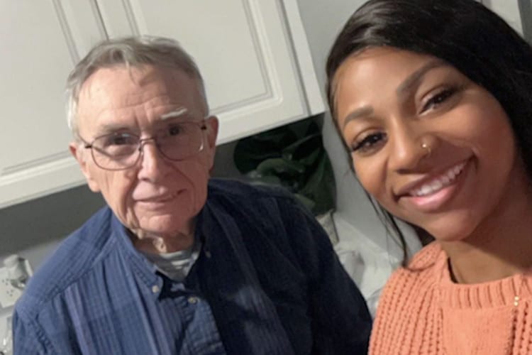 A selfie featuring a young woman smiling brightly next to an older man with glasses and gray hair, who looks directly at the…