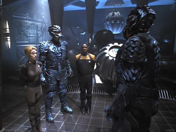 A still from Star Trek: Voyager showing four characters in a dimly lit, futuristic room with metallic walls and a large…