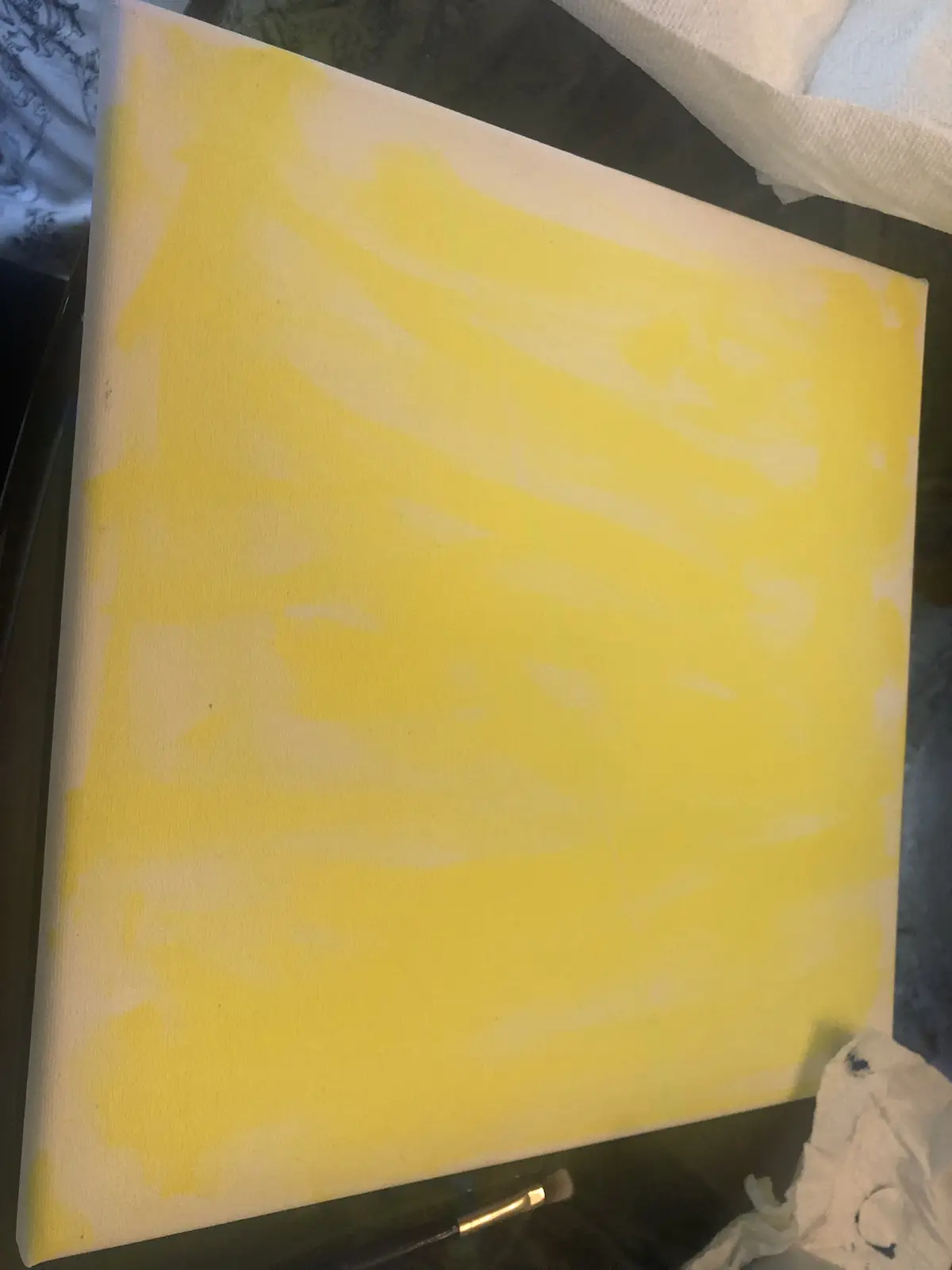 A close-up photo shows a square canvas with a rough, uneven layer of yellow paint applied over a white background, with…