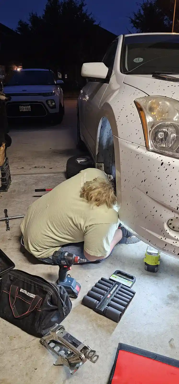 A person with blonde hair is kneeling beside a white car with its front wheel removed and body covered in black speckles…
