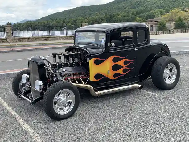 A black classic hot rod with a large exposed engine, chrome wheels, and orange flame decals on its side is parked in a lot…