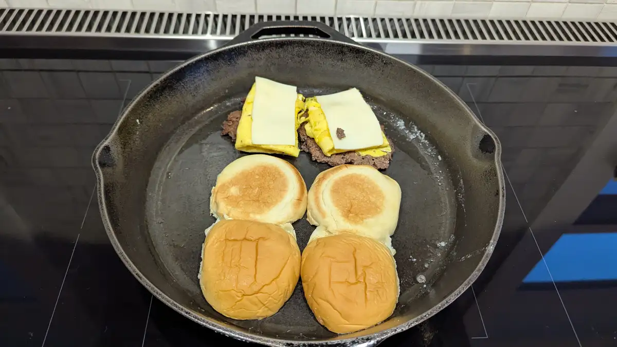 A cast iron skillet on a stovetop contains two breakfast sliders being cooked: each has a patty, scrambled egg, and cheese…