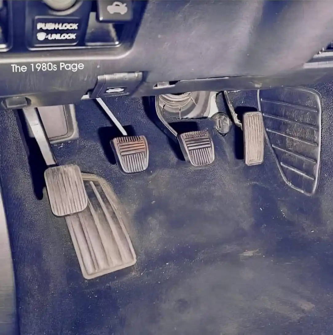 A close-up photo of a car's worn, dusty floor pedals showing the clutch, brake, and accelerator, with a 'PUSH LOCK -UNLOCK'…