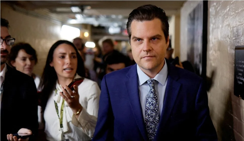 A man in a blue suit and patterned tie stares intensely forward in a hallway, surrounded by blurred people. A woman behind…