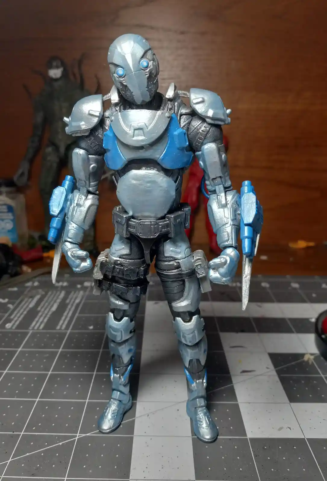 A custom-painted action figure of a humanoid robot with silver and blue armor, glowing blue eyes, and dual blades, standing…