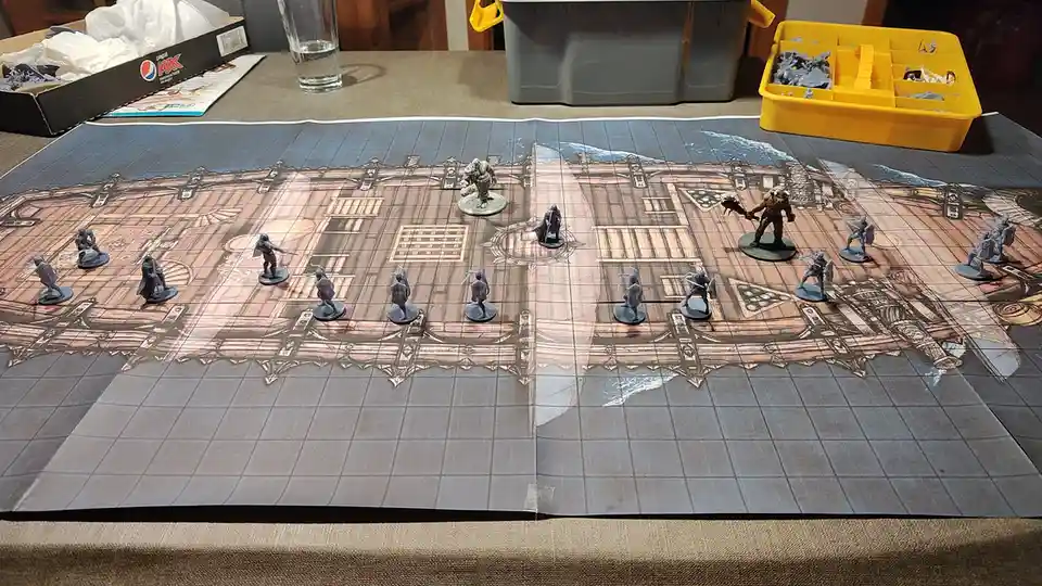 A tabletop miniature wargaming scene shows two opposing armies of grey miniatures facing each other on a printed battle map…
