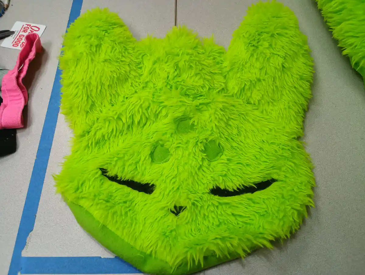 A bright neon green, fluffy, bunny-shaped plush mask with stitched black eyes and a smile lies on a gray surface, partially…