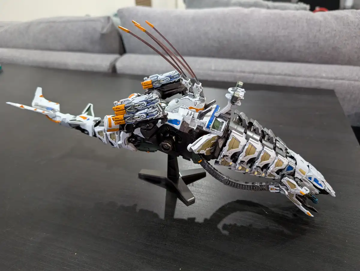 A highly detailed, white, blue, and gold robot model shaped like a shark is displayed on a black stand on a dark table, with…