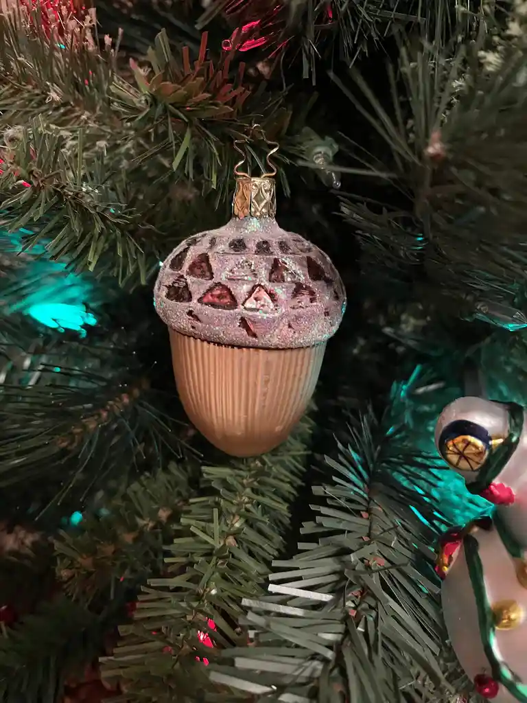 A close-up of a Christmas tree ornament shaped like an acorn, with a glittery pink cap and a tan body, hanging among green…