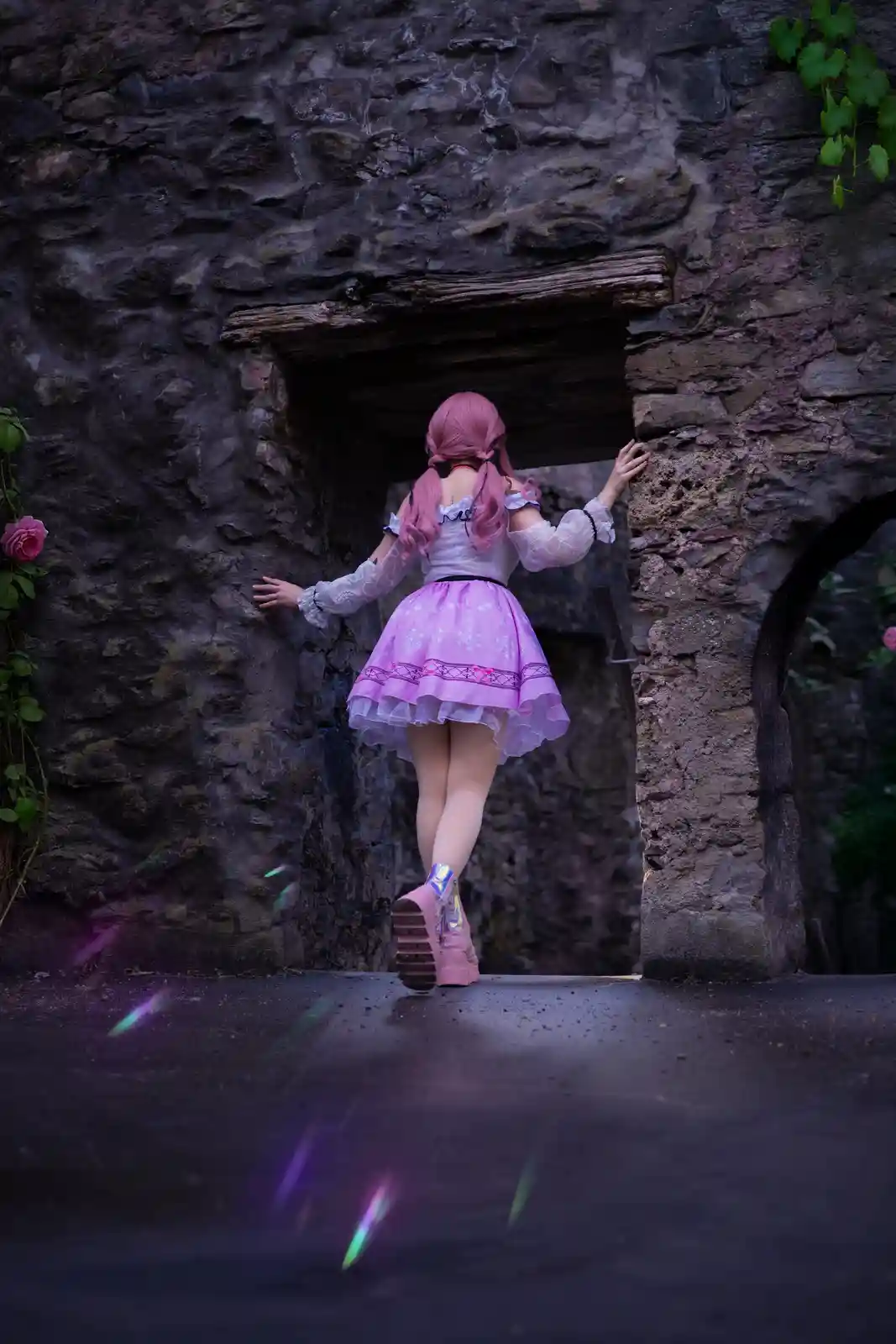 A person with pink hair and a pink and white frilly outfit stands with their back to the camera, stepping through a dark…
