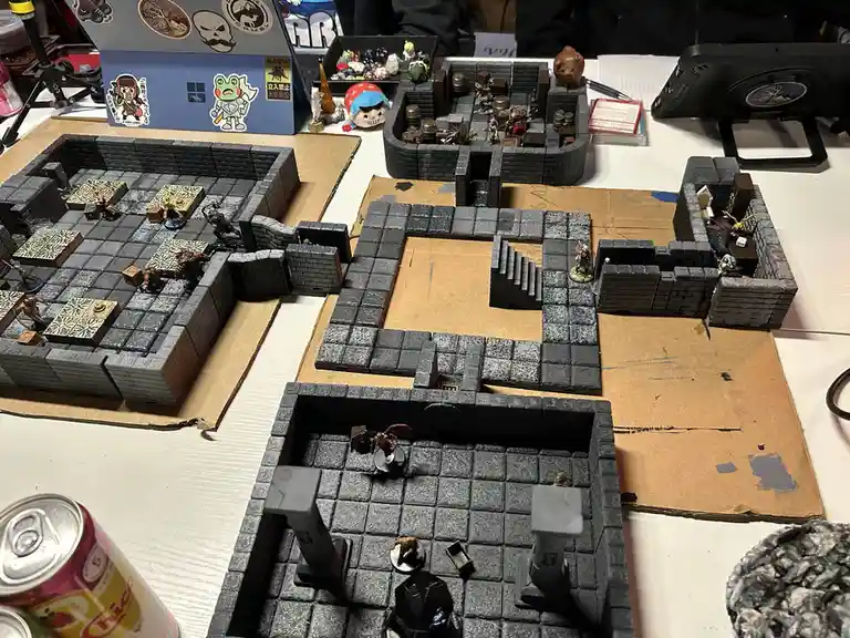 A tabletop role-playing game setup featuring multiple interconnected dungeon rooms made of gray brick-like tiles on…