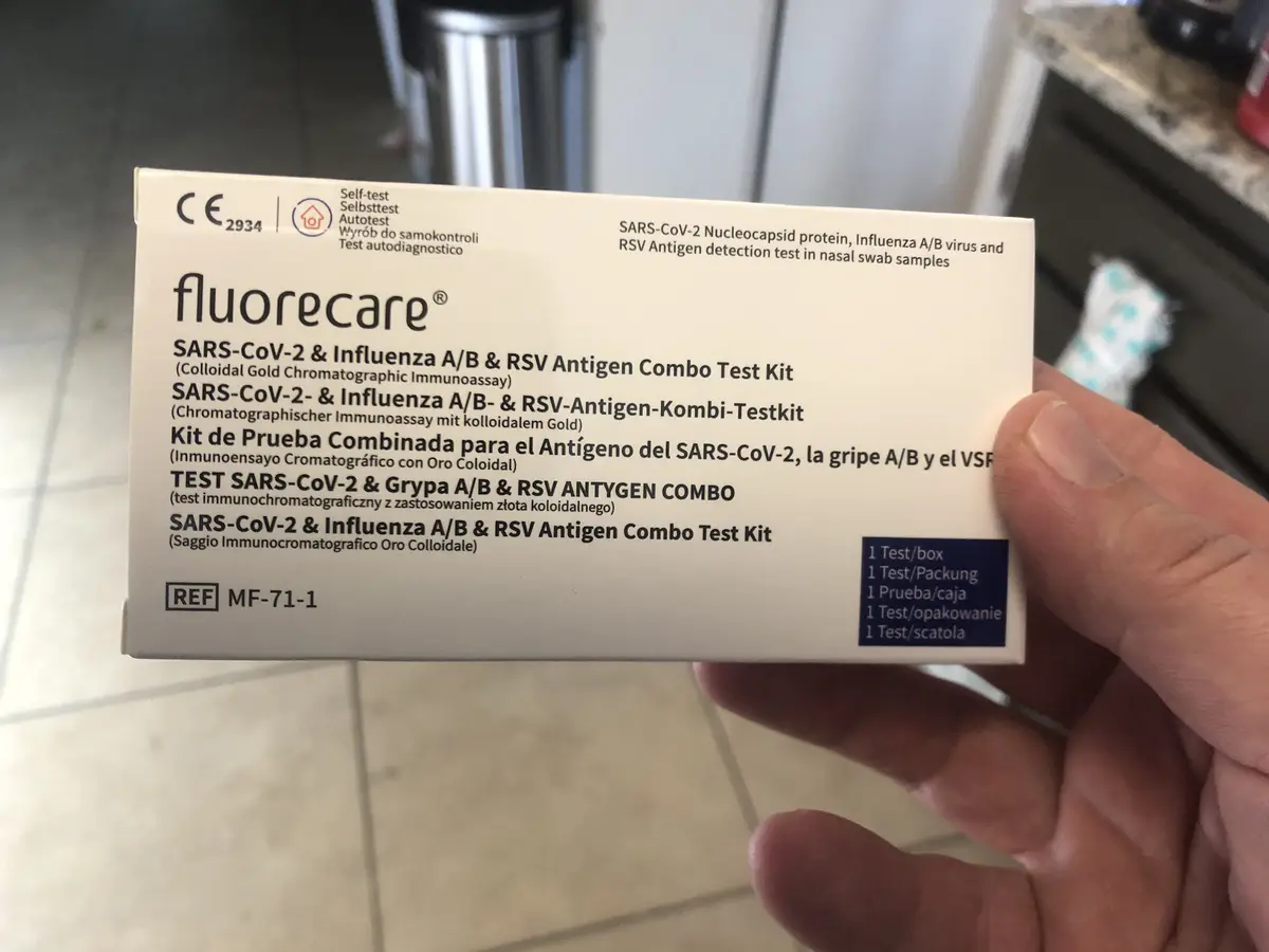 A hand holds a white box labeled 'fluorecare' which is a SARS-CoV-2 & Influenza A/B & RSV Antigen Combo Test Kit. The box…