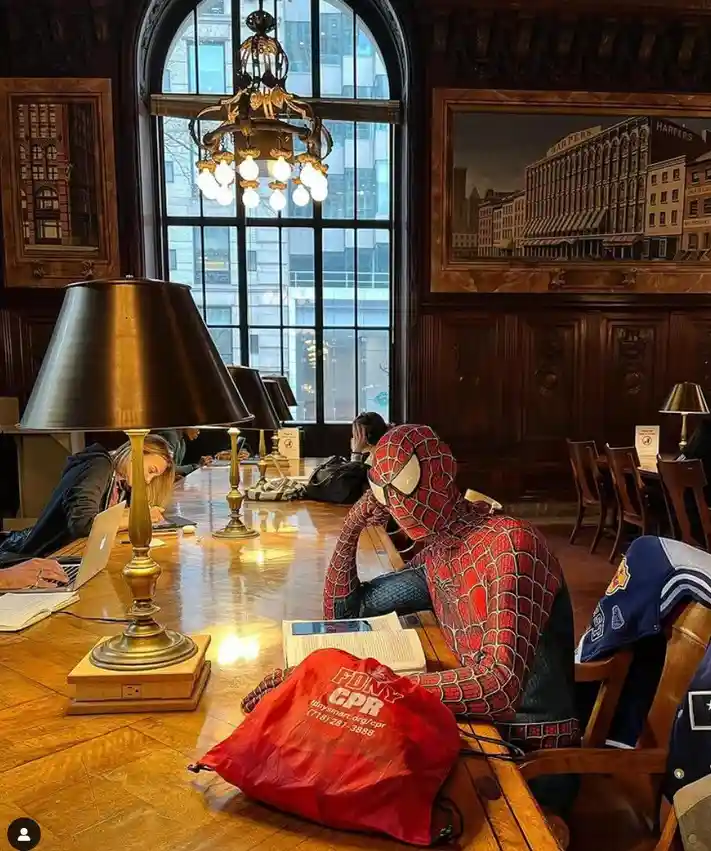 A person in a full Spider-Man costume sits at a wooden library table, appearing to study with an open book and tablet. A red…