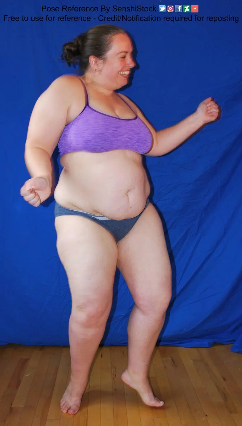 A woman in a purple sports bra and grey bikini bottoms stands barefoot on a wooden floor in front of a blue backdrop…