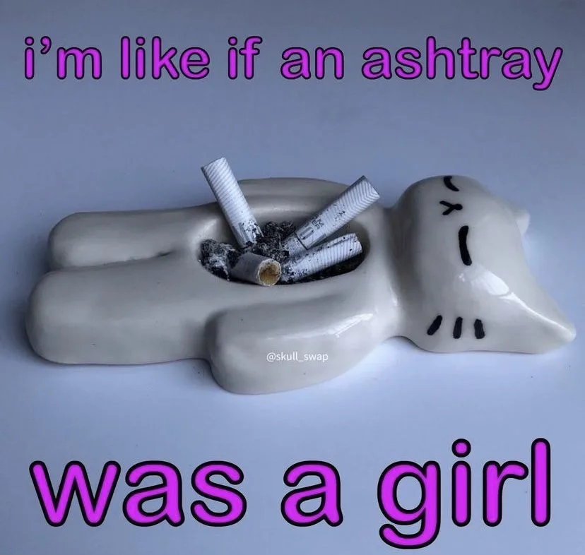 A ceramic ashtray shaped like a sleeping girl with a simple face drawn on her head, filled with cigarette butts and ash. The…