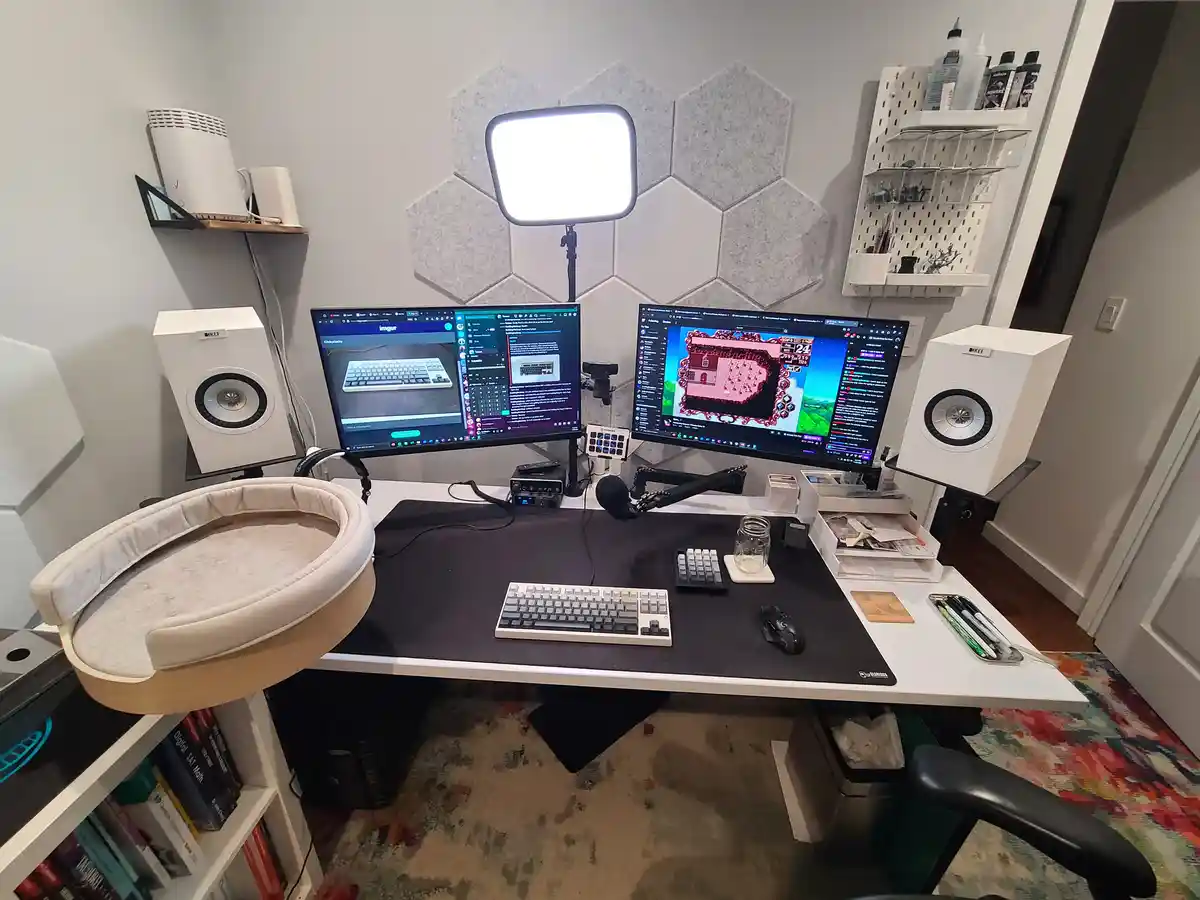 A well-organized home office desk with two monitors, a white keyboard, and professional audio equipment, including two white…
