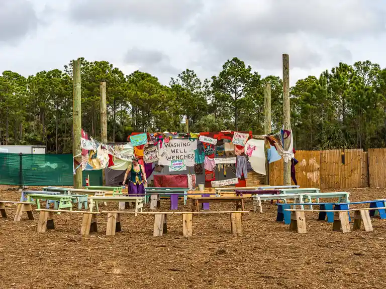 An outdoor medieval-themed laundry setup with colorful wooden benches arranged in rows on mulch ground, clothes hanging on…