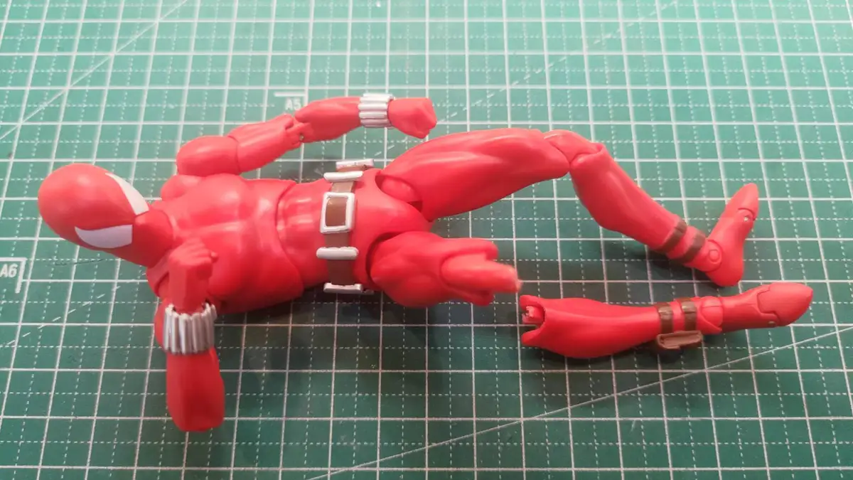 A red Spider-Man action figure lies on a green cutting mat with its legs splayed out, one leg detached and lying nearby. The…
