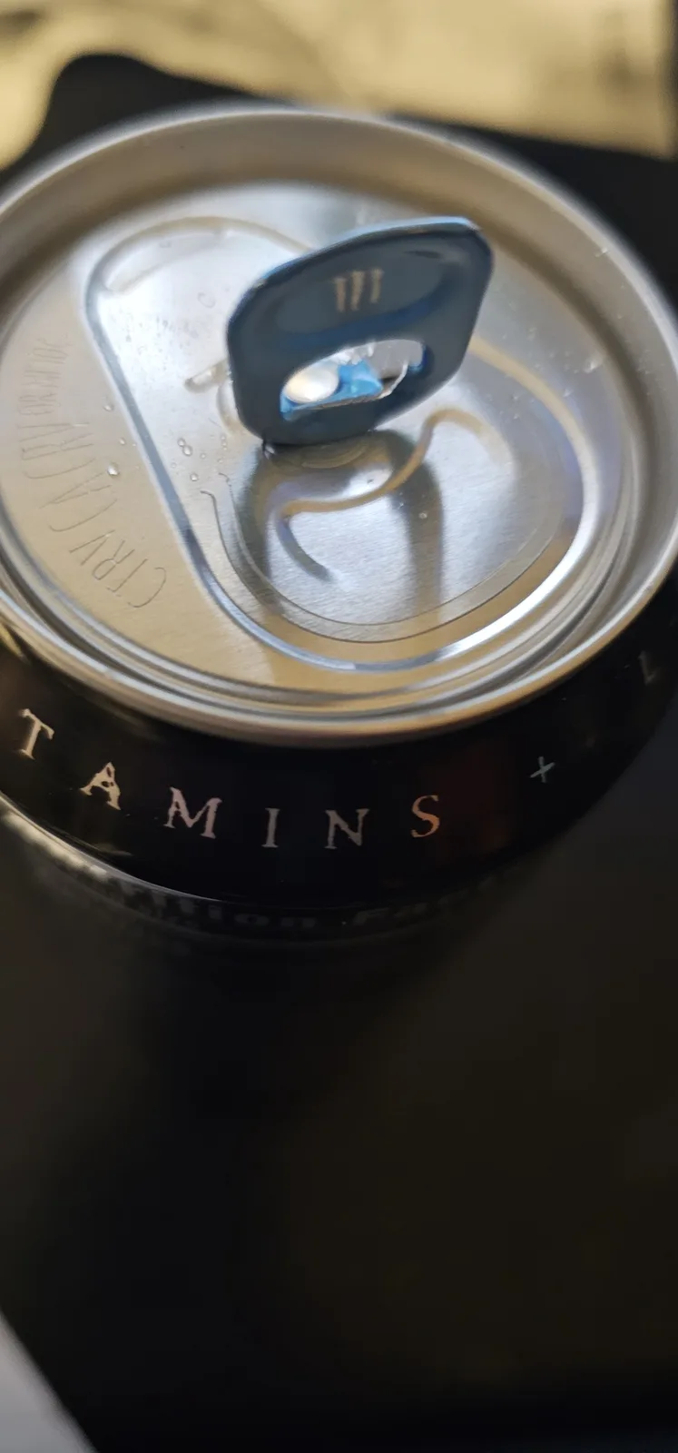 A close-up photo of a black beverage can with 'TAMINS' printed in gold lettering on its side, showing a partially opened…