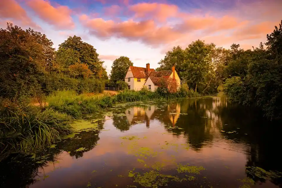 A picturesque countryside scene shows a white cottage with a red-tiled roof nestled beside a calm river at sunset…