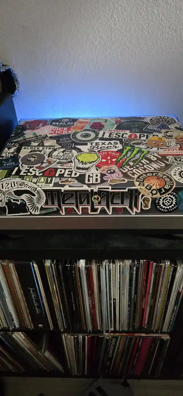 A close-up of a black shelf covered in numerous colorful stickers, including a large 'Metalachi' logo and 'I ESCAPED GB'…