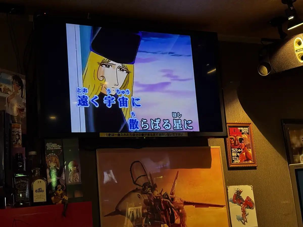 A TV screen in a dimly lit room displays an anime scene with a blonde-haired character wearing a black hat against a purple…