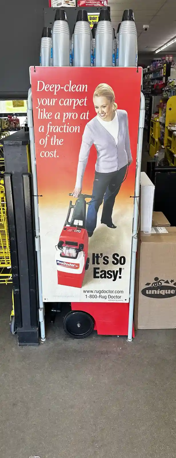A red Rug Doctor carpet cleaning machine display in a store, featuring an advertisement with a smiling woman pushing the…