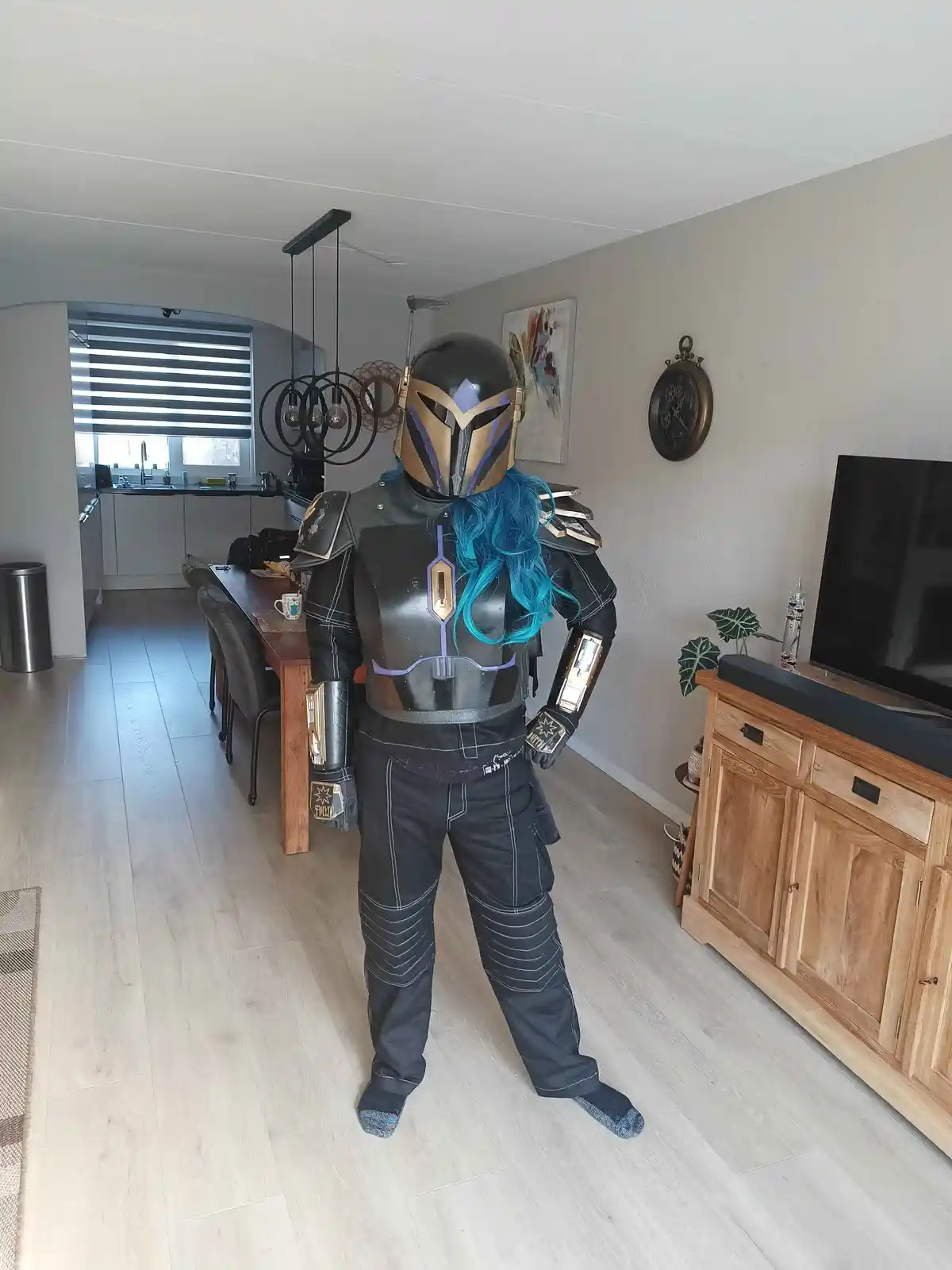 A person in a Mandalorian-style costume with a black helmet and armor stands in a modern home, but has a bright blue wig…