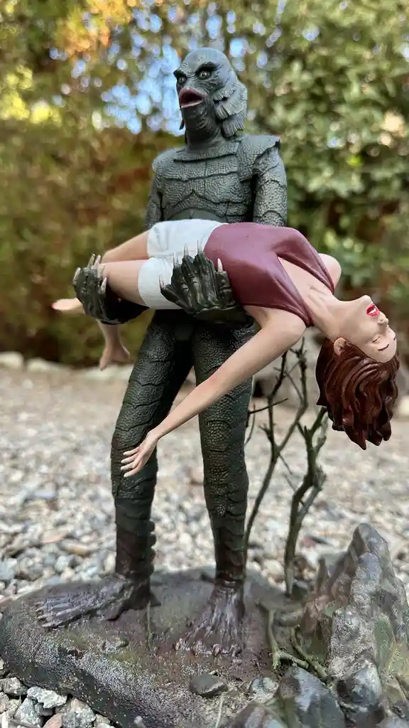 A figurine of the Creature from the Black Lagoon stands outdoors holding a woman upside down, her head pointing toward the…