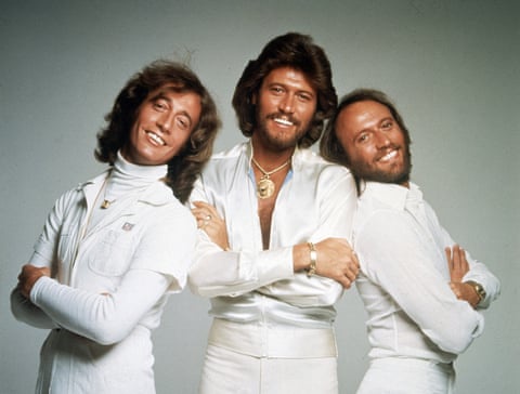 Three men in matching white 1970s-style outfits pose confidently with arms crossed against a plain gray background. The man…
