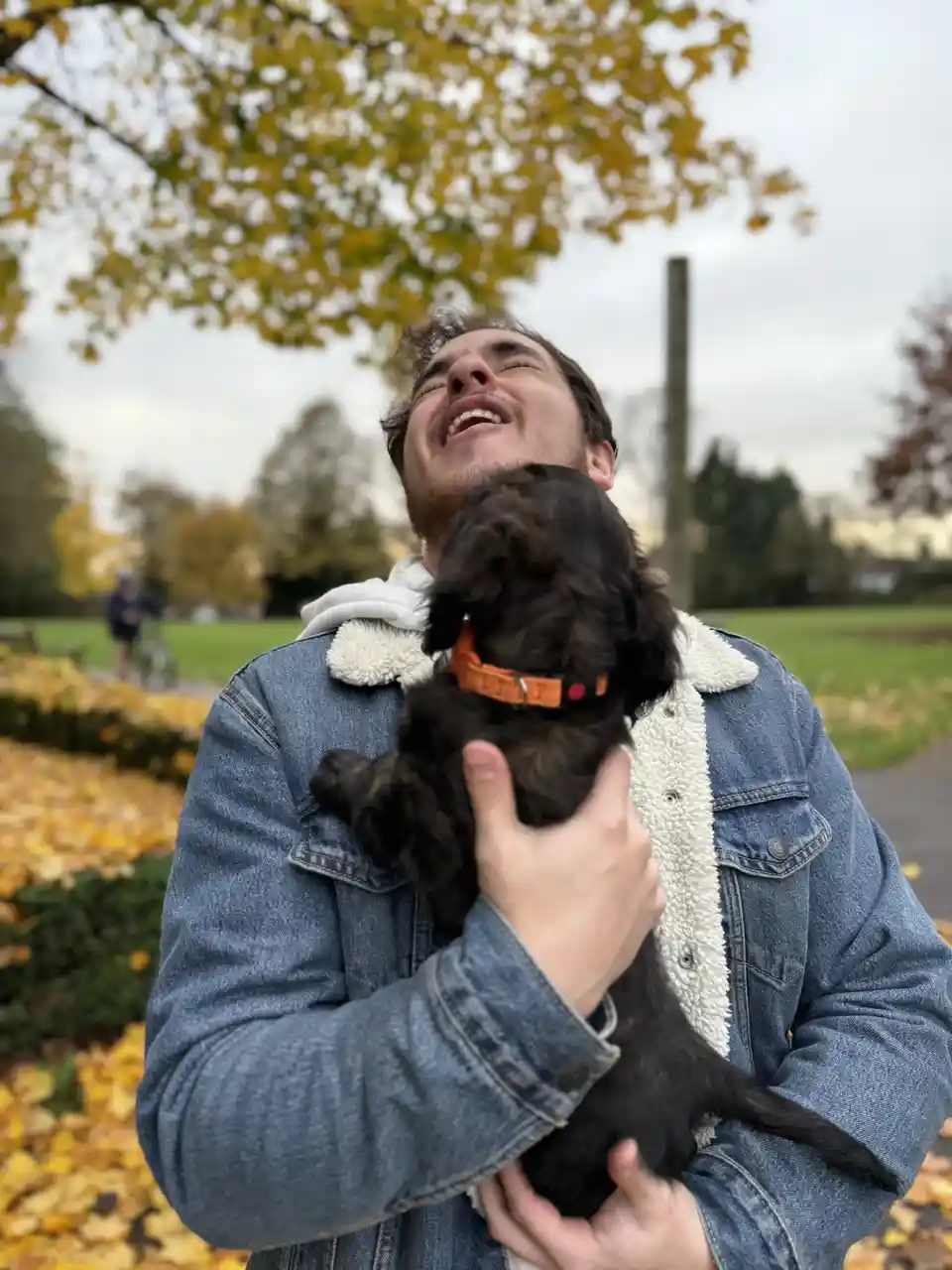 A man in a denim jacket with a white fleece collar is holding a small black puppy with an orange collar. He is laughing with…