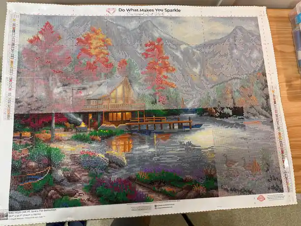 A partially completed diamond painting by Diamond Art Club titled 'Space For Reflection' shows a scenic lakeside cabin with…