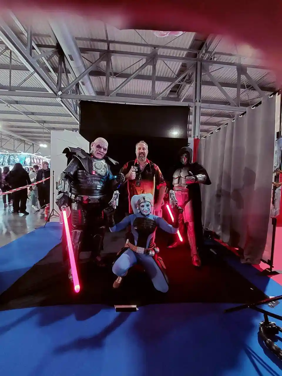 Four people pose together in a photo booth at a convention, three dressed as Star Wars characters holding glowing red…