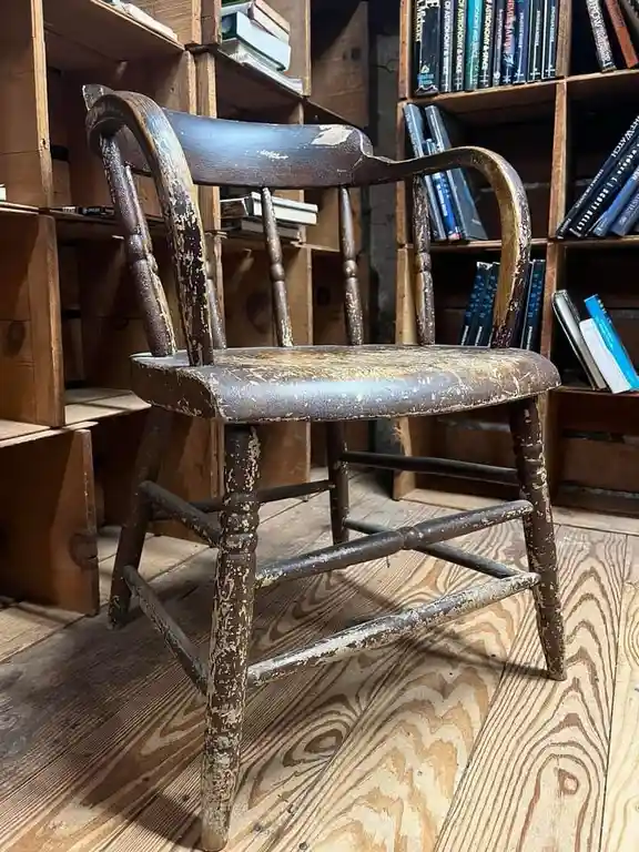 a heavily distressed, dark wooden armchair with peeling paint and worn wood sits on a light-colored wooden floor, surrounded…