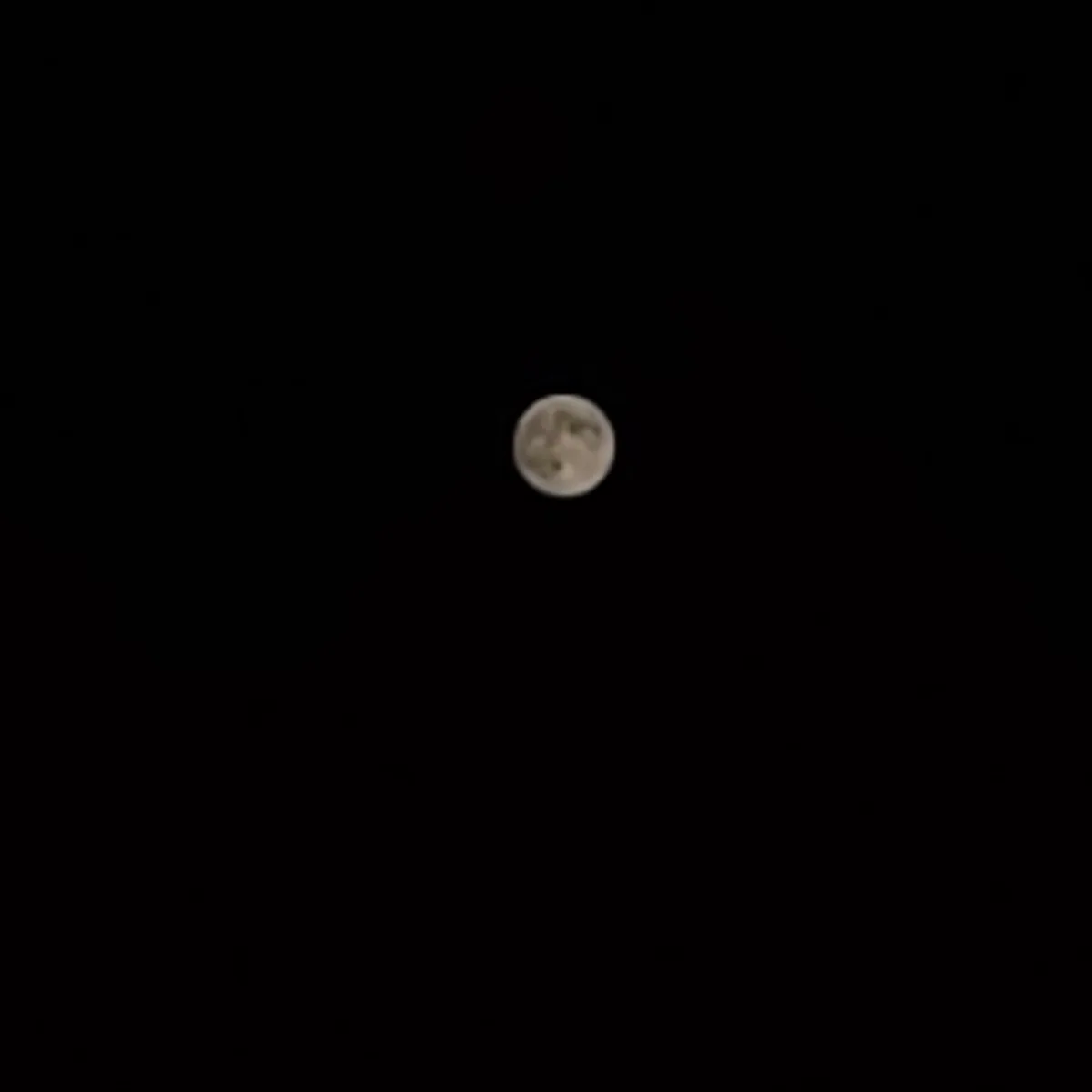 A blurry, low-resolution image shows a pale, round moon centered against a completely black sky. The moon's surface features…