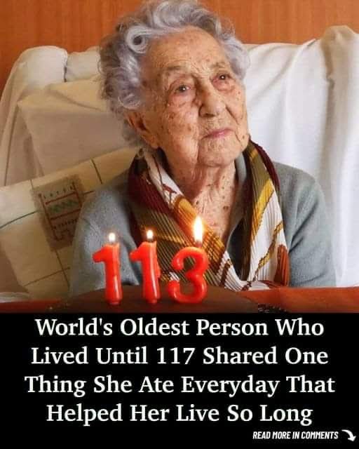 An elderly woman with gray hair sits in a chair, looking toward the viewer. In front of her on a table are three red candles…