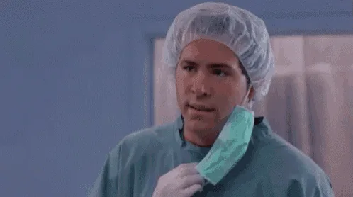 A man in surgical scrubs and a hairnet holds a surgical mask, looking confused and increasingly bewildered as the frames…
