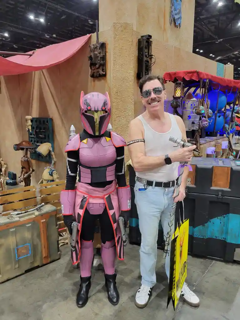 A man in a white tank top and sunglasses stands next to a person in a full-body pink and black Mandalorian-style armor…