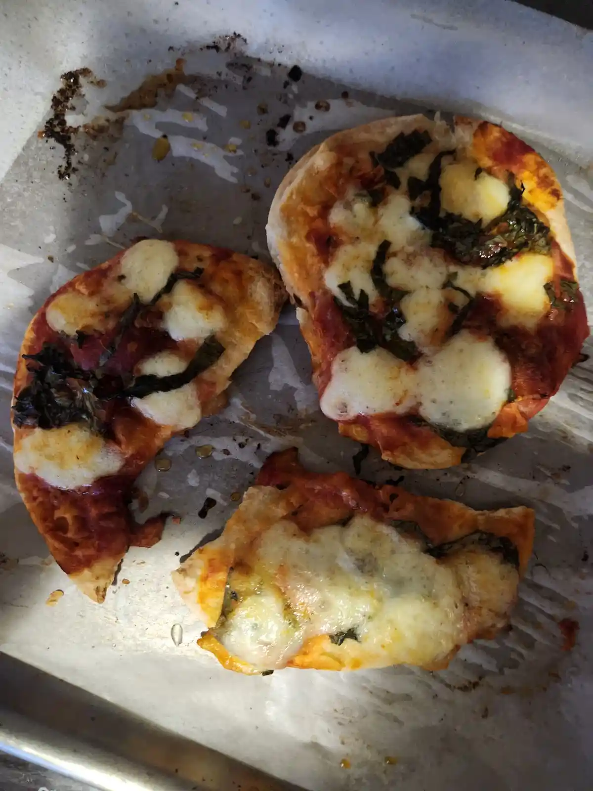 Three small, round, bagel-like bases topped with tomato sauce, melted cheese, and wilted basil leaves, arranged on a baking…