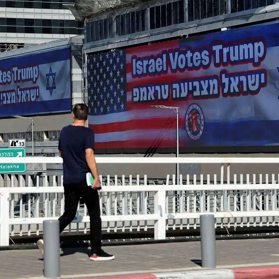 A man walks away from the camera on a sidewalk in front of large banners. The banners feature the American flag and text in…