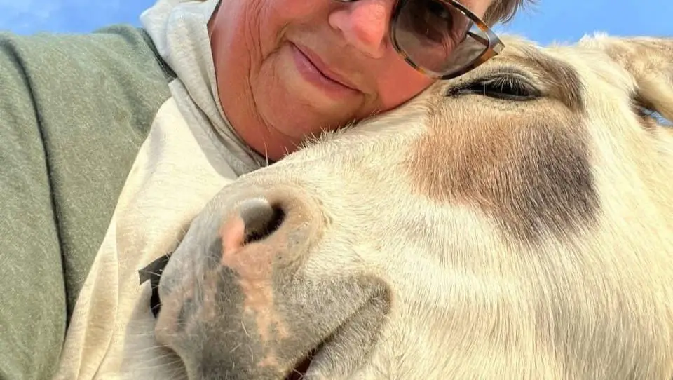 A close-up selfie shows a smiling person wearing sunglasses and a hoodie, resting their head affectionately against the…