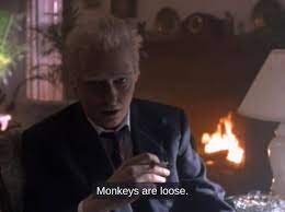A man with white hair and a dark suit sits in a dimly lit room with a fireplace behind him, looking alarmed. He holds a…