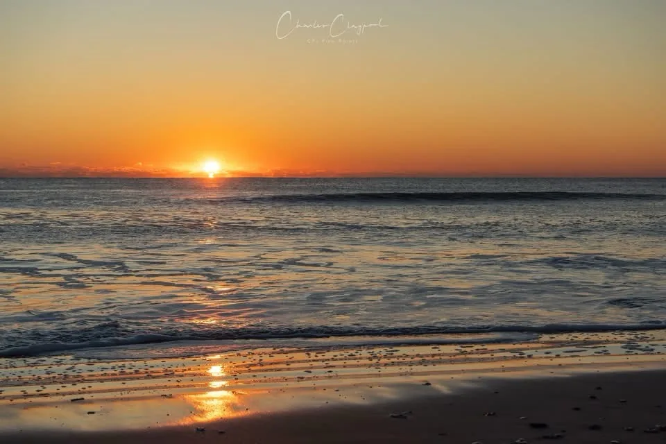 A serene sunset over the ocean with the sun low on the horizon, casting orange and yellow light across the water and wet…