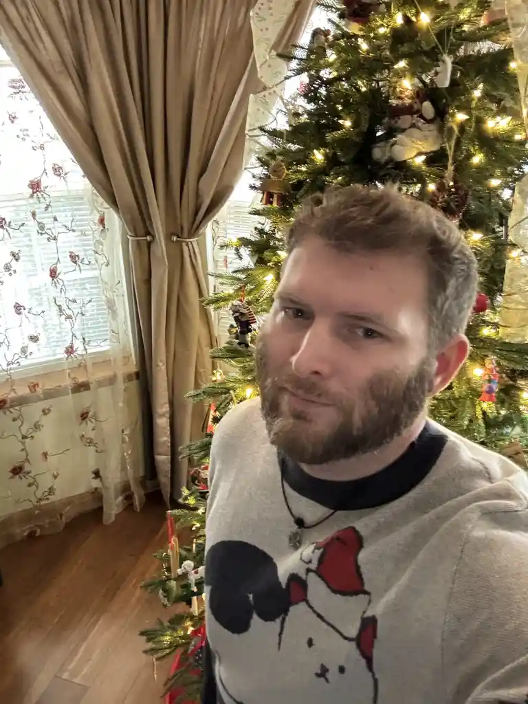 A man with a beard and a festive sweater featuring a cartoon character stands in front of a decorated Christmas tree with…